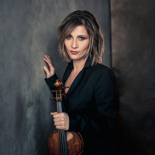 A white woman with shoulder length brown hair leans against a gray painted wall holding her violin while wearing a dark suit jacket. 