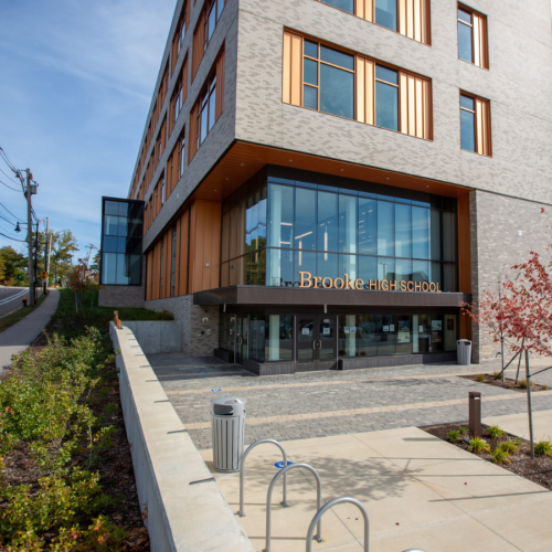 A lowrise contemporary school building, clad in light gray stone, with orange windowframes. A glass atrium with Brooke High School over the doors.