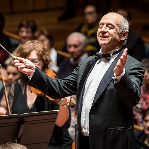 An older white man in a suit and a bow tie conducts an orchestra with arms outstretched in front of a crowd. 
