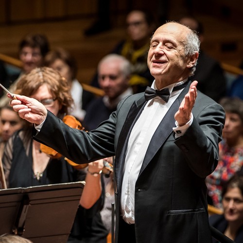 An older white man in a suit and a bow tie conducts an orchestra with arms outstretched in front of a crowd. 