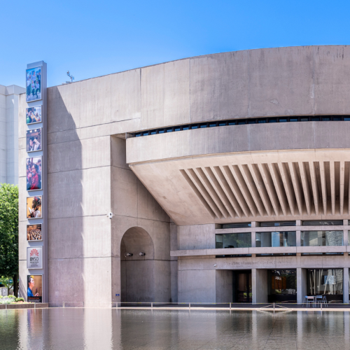 the curved facade of a modern-style building, with a glass atrium at street level and a tower with stacked photos rising to the left