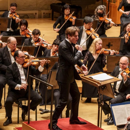 A young man conducts a large orchestra on a red podium; behind him the violin section is visible in formal concert attire. 