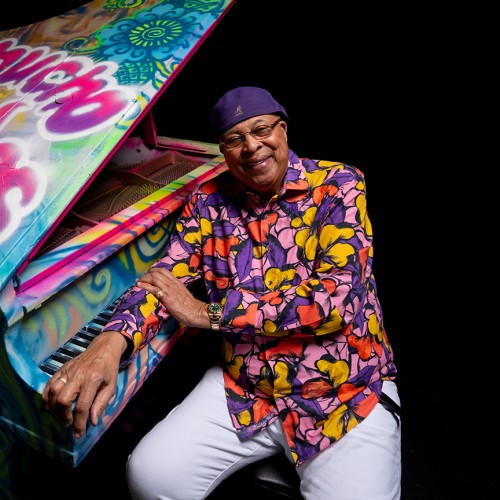 An older black man in a colorful purple, pink, and yellow top and a purple cap leans on a grand piano covered in colorful graffiti. 