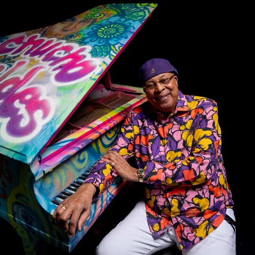 An older black man in a colorful purple, pink, and yellow top and a purple cap leans on a grand piano covered in colorful graffiti. 