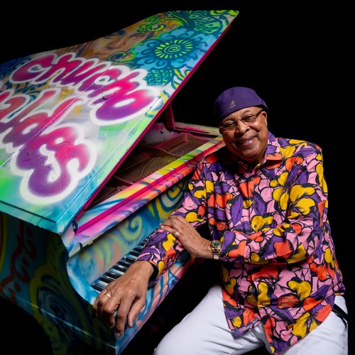 Chucho Valdes, an older Afro-Latin man in a colorful purple, pink, and yellow top and a purple cap leans on a grand piano covered in colorful graffiti. 