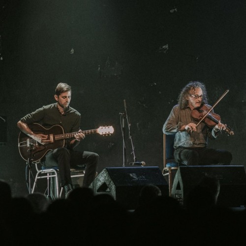 an ensemble of folk musicians play stringed instruments under white light on a dark stage