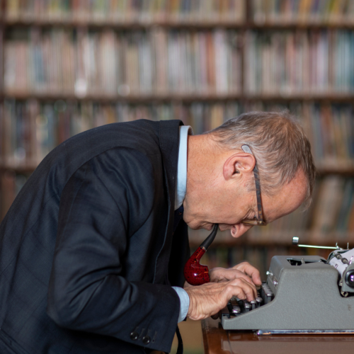 A white man in a jacket and glasses with pipe a dangling out of his mouth leans over a grey typewriter in front of a bookcase. 