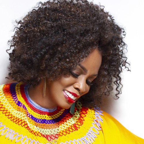 A black woman looking downward, shoulder length curly hair, wearing gold earrings and a bright yellow shirt with a colorful collar 