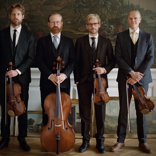 Four white men in a string quartet stand in a row in dark suits holding their instruments in front of several nature murals. 