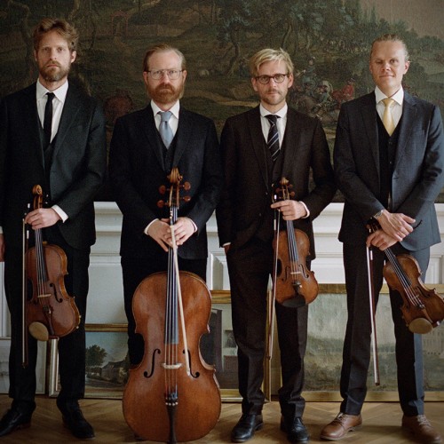 Four white men in a string quartet stand in a row in dark suits holding their instruments in front of several nature murals. 