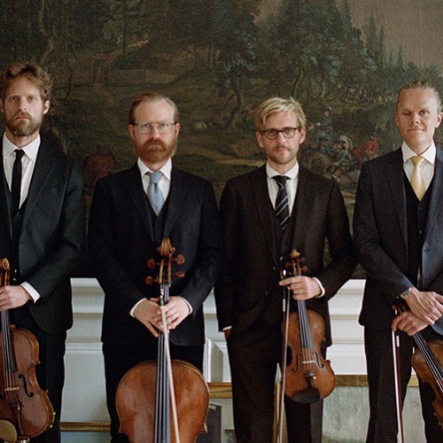 Four white men in a string quartet stand in a row in dark suits holding their instruments in front of several nature murals. 