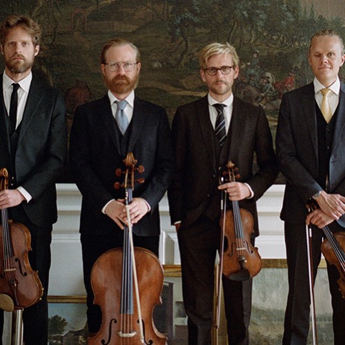 Four white men in a string quartet stand in a row in dark suits holding their instruments in front of several nature murals. 