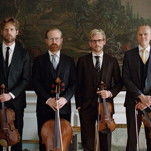 Four white men in a string quartet stand in a row in dark suits holding their instruments in front of several nature murals. 