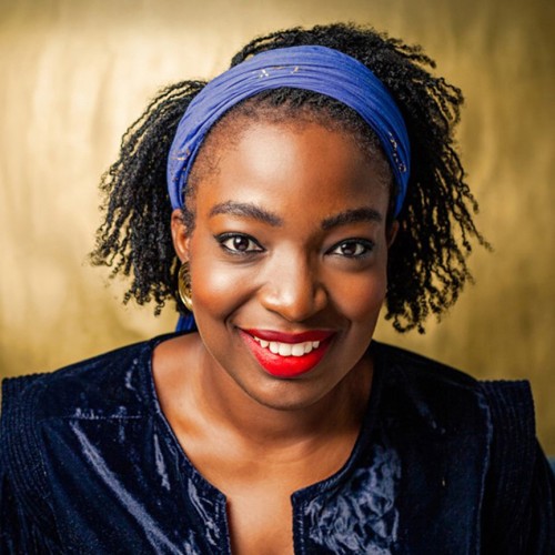 A smiling black woman with a blue headband, red lipstick, gold earrings, and a velvet like dark blue jacket.