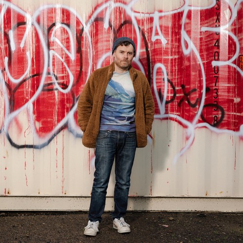 An American, white man stands in blue jeans, a t-shirt with mountains, and a brown fuzzy jacket in front of a wall of graffiti 