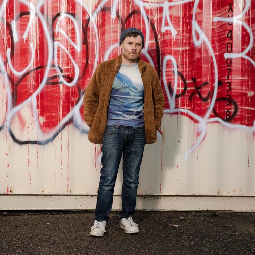 An American, white man stands in blue jeans, a t-shirt with mountains, and a brown fuzzy jacket in front of a wall of graffiti 