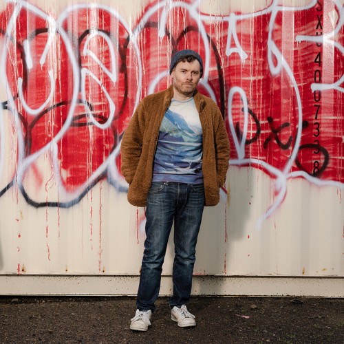 An American, white man stands in blue jeans, a t-shirt with mountains, and a brown fuzzy jacket in front of a wall of graffiti 