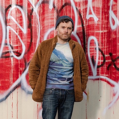 An American, white man stands in blue jeans, a t-shirt with mountains, and a brown fuzzy jacket in front of a wall of graffiti 