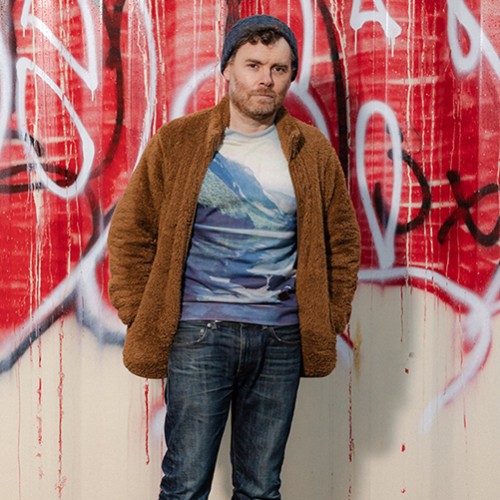 An American, white man stands in blue jeans, a t-shirt with mountains, and a brown fuzzy jacket in front of a wall of graffiti 