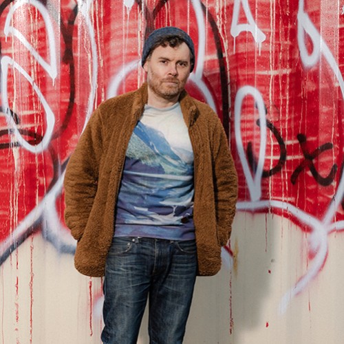 An American, white man stands in blue jeans, a t-shirt with mountains, and a brown fuzzy jacket in front of a wall of graffiti 
