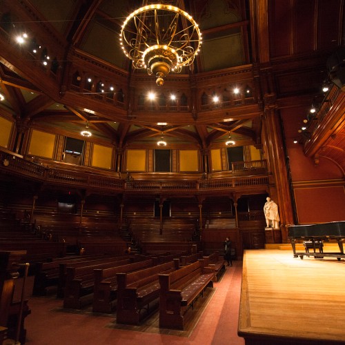 A wood paneled auditorium with a bright chandelier and golden lights illuminating dark wooden pews