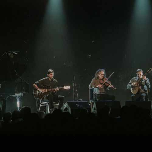 Five musicians are lined up in front of a large crowd playing a variety of instruments with two spotlights shining down on them. 