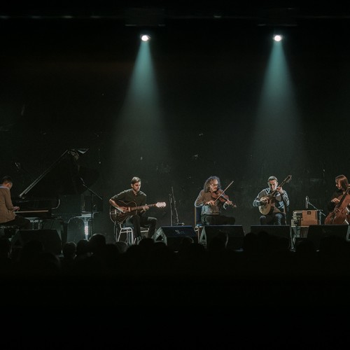 Five musicians are lined up in front of a large crowd playing a variety of instruments with two spotlights shining down on them.