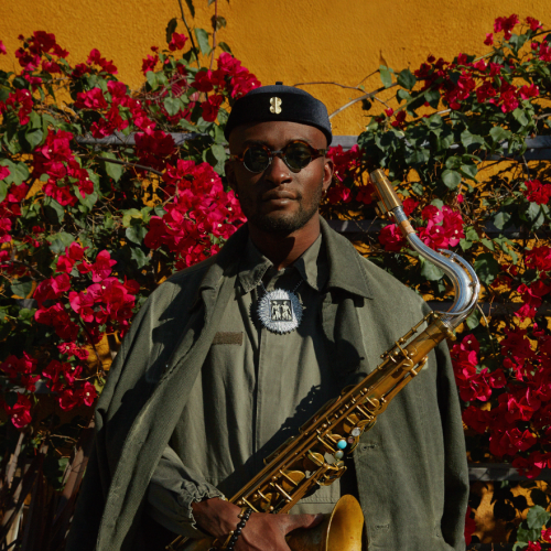 Isaiah Collier wears sunglasses and a military inspired cape and jumpsuit under warm light in an outdoor garden