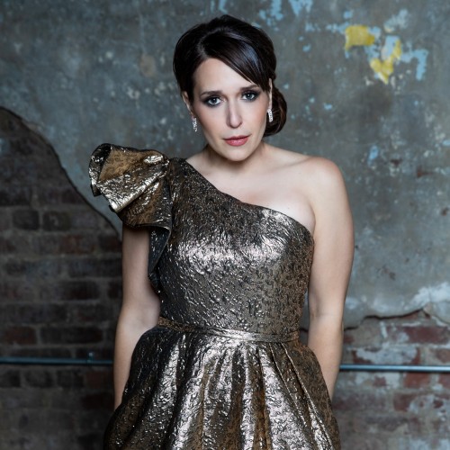 a woman with pale skin and dark hair wears a one-shoulder crinkly textured metallic fabric dress. her chin slightly tipped down, she makes intense eye contact.