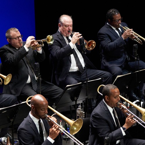 Four members of a jazz band are shown playing brass instruments in suits with polka dot ties on a stage with a blue background 