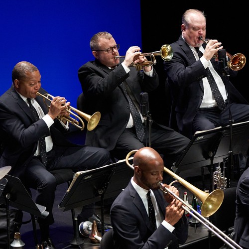 Four members of a jazz band are shown playing brass instruments in suits with polka dot ties on a stage with a blue background 