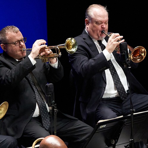 Four members of a jazz band are shown playing brass instruments in suits with polka dot ties on a stage with a blue background 