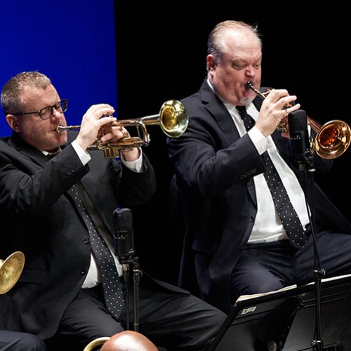 Four members of a jazz band are shown playing brass instruments in suits with polka dot ties on a stage with a blue background 