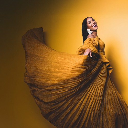 a black woman, in golden light, wearing a golden dress, turns and gestures to us with one arm as she glides forward