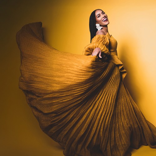 A black woman flourishes a pleated yellow dress while smiling widely in front of a shadowy yellow background. 