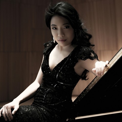 A South Korean woman in a black sparkly dress leans on a grand piano lid, her dark curly hair cascading. 