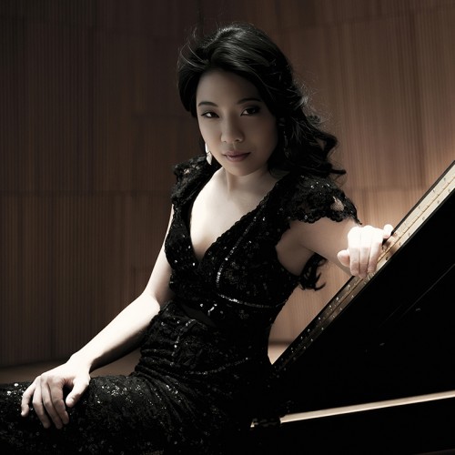A South Korean woman in a black sparkly dress leans on a grand piano lid, her dark curly hair cascading. 
