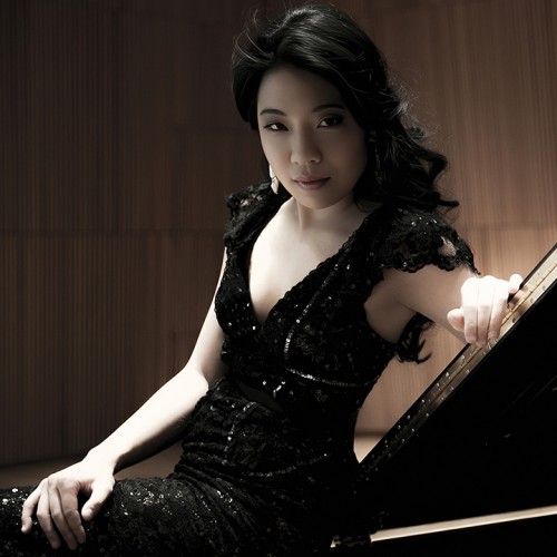 A South Korean woman in a black sparkly dress leans on a grand piano lid, her dark curly hair cascading. 
