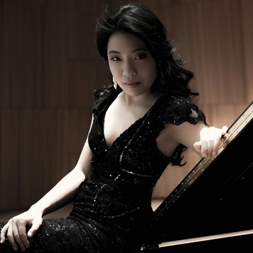 A South Korean woman in a black sparkly dress leans on a grand piano lid, her dark curly hair cascading. 