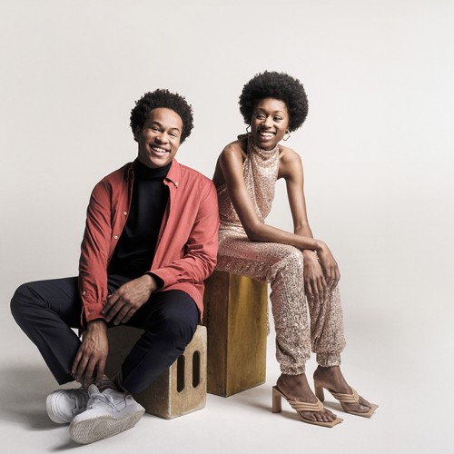 sheku and isata kanneh-mason, english siblings of caribbean heritage, smile. she wears a light tan sleeveless jumpsuit and he wears an open pink shirt over a dark t-shirt and dark trousers