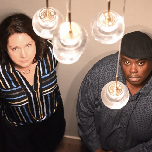 A white woman stands between two black men; the trio looks up with stoic expressions towards a lighting fixture with clear globes. 