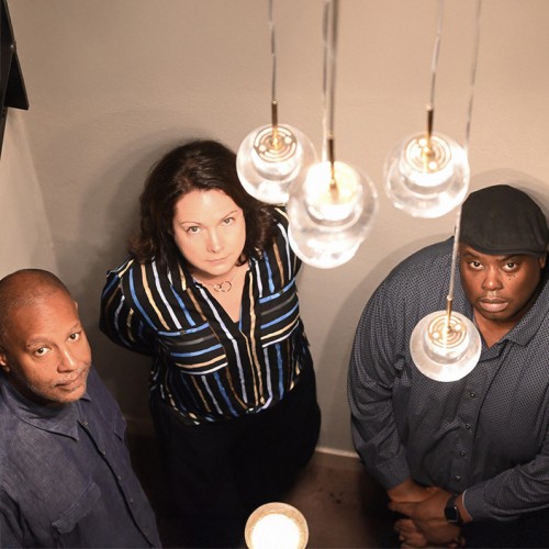 A white woman stands between two black men; the trio looks up with stoic expressions towards a lighting fixture with clear globes. 