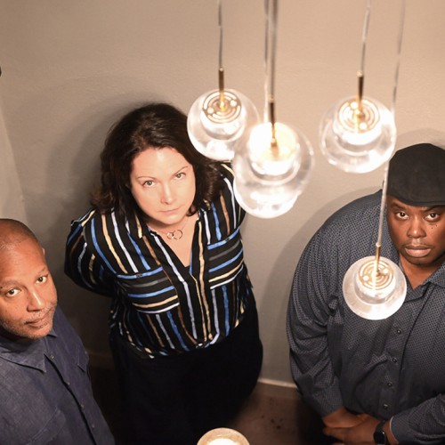 A white woman stands between two black men; the trio looks up with stoic expressions towards a lighting fixture with clear globes. 