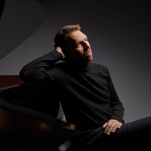 A white, Norwegian man in a black sweat sits next to a grand piano, leaning one arm against the curve of the piano 