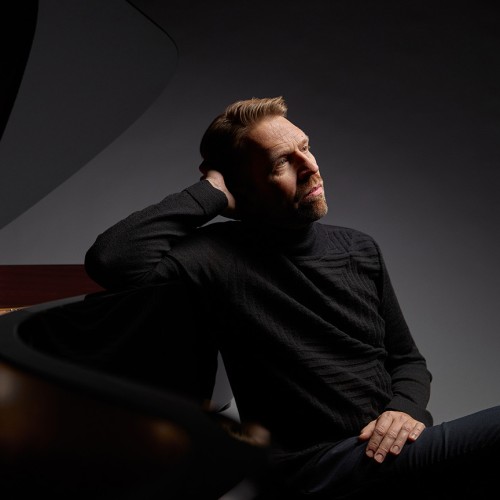 A white, Norwegian man in a black sweat sits next to a grand piano, leaning one arm against the curve of the piano 