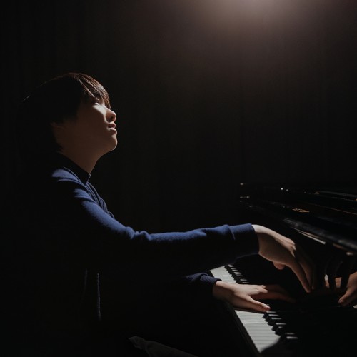 A young, Japanese man sits playing the piano as a spotlight shines from above. 