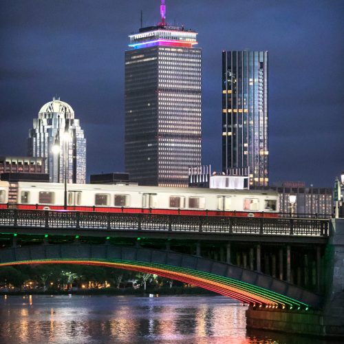 Boston's red line train cars cross the longfellow bridge. the bridge and the prudential center behind it are illuminated with Pride rainbow colors