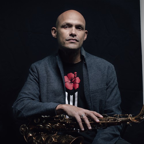 A Puerto Rican man in a gray jacket sits with a saxophone across his lap in front of a black background. 