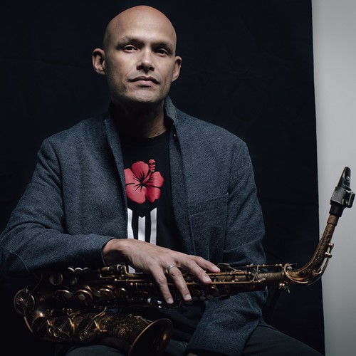A Puerto Rican man in a gray jacket sits with a saxophone across his lap in front of a black background. 