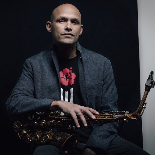 A Puerto Rican man in a gray jacket sits with a saxophone across his lap in front of a black background. 